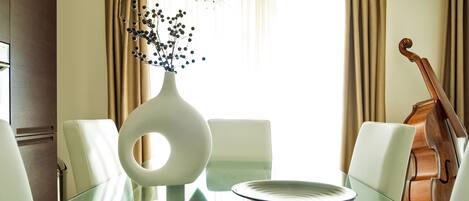 En spiseplads med et glasbord, hvide stole, en vase med blomster og en cello i baggrunden.
