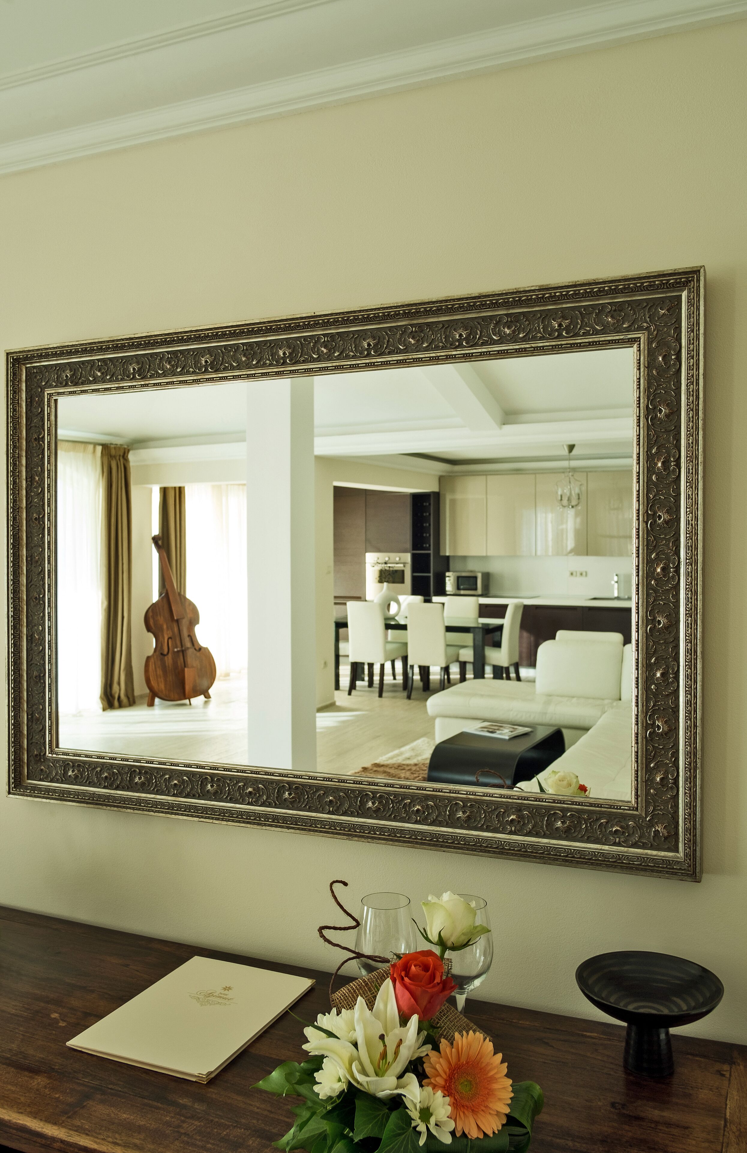 A mirror with an ornate frame reflecting a living room with a dining area, a cello, and a vase with flowers.