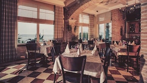 A restaurant with a checkered floor, brick walls, and large windows overlooking the sea. Tables are set with white tablecloths and napkins. - Hotel Azimut (Sveti Stefan)