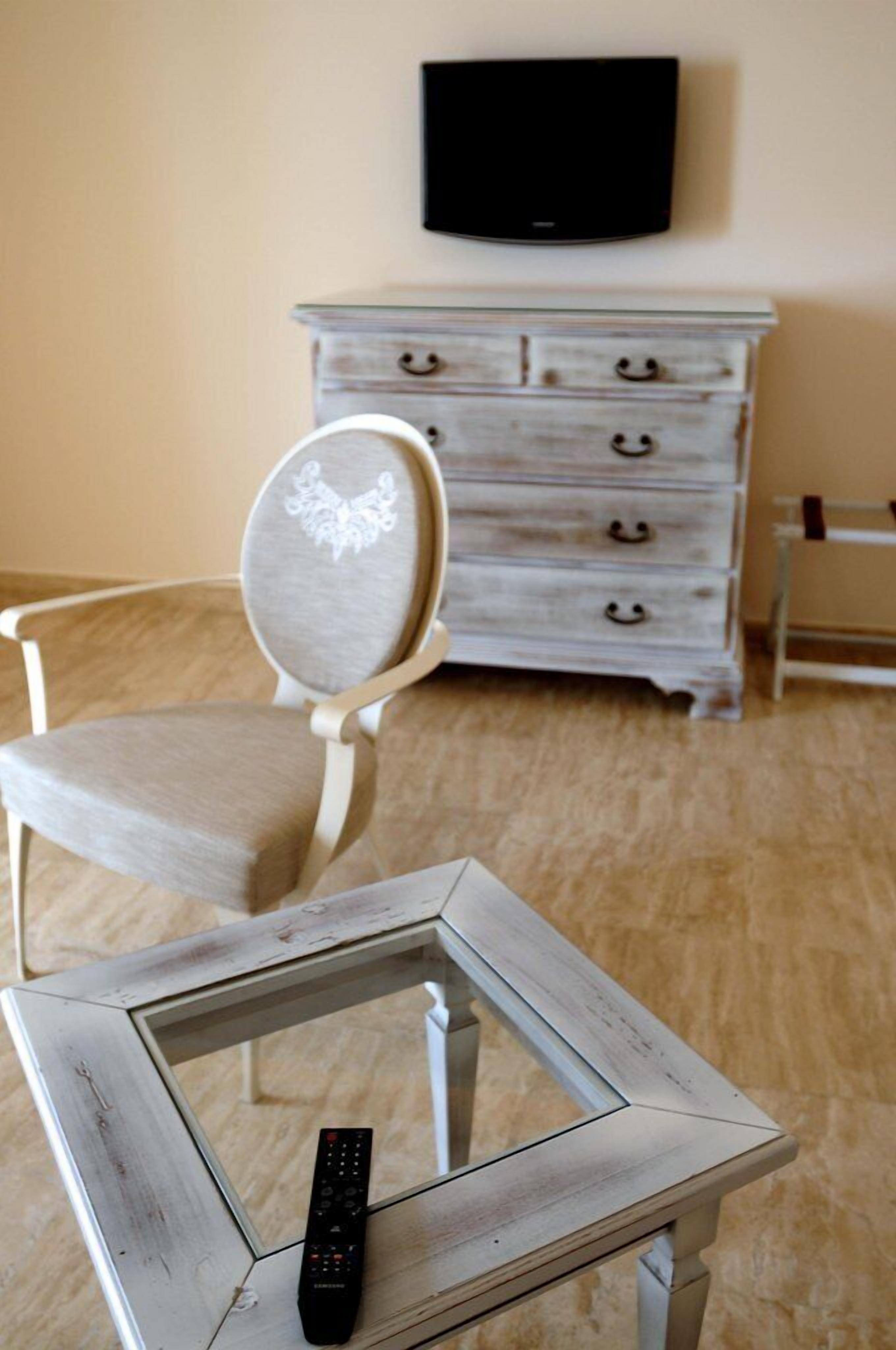 Una habitación con una mesa de marco blanco, una silla con cojín blanco, un televisor montado en la pared y un aparador blanco con cajones.