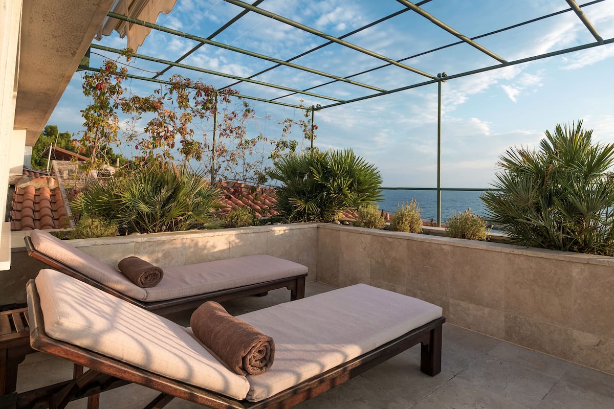 Un espace de détente extérieur baigné de soleil, avec deux fauteuils en bois, un mur en béton et une vue sur la mer.