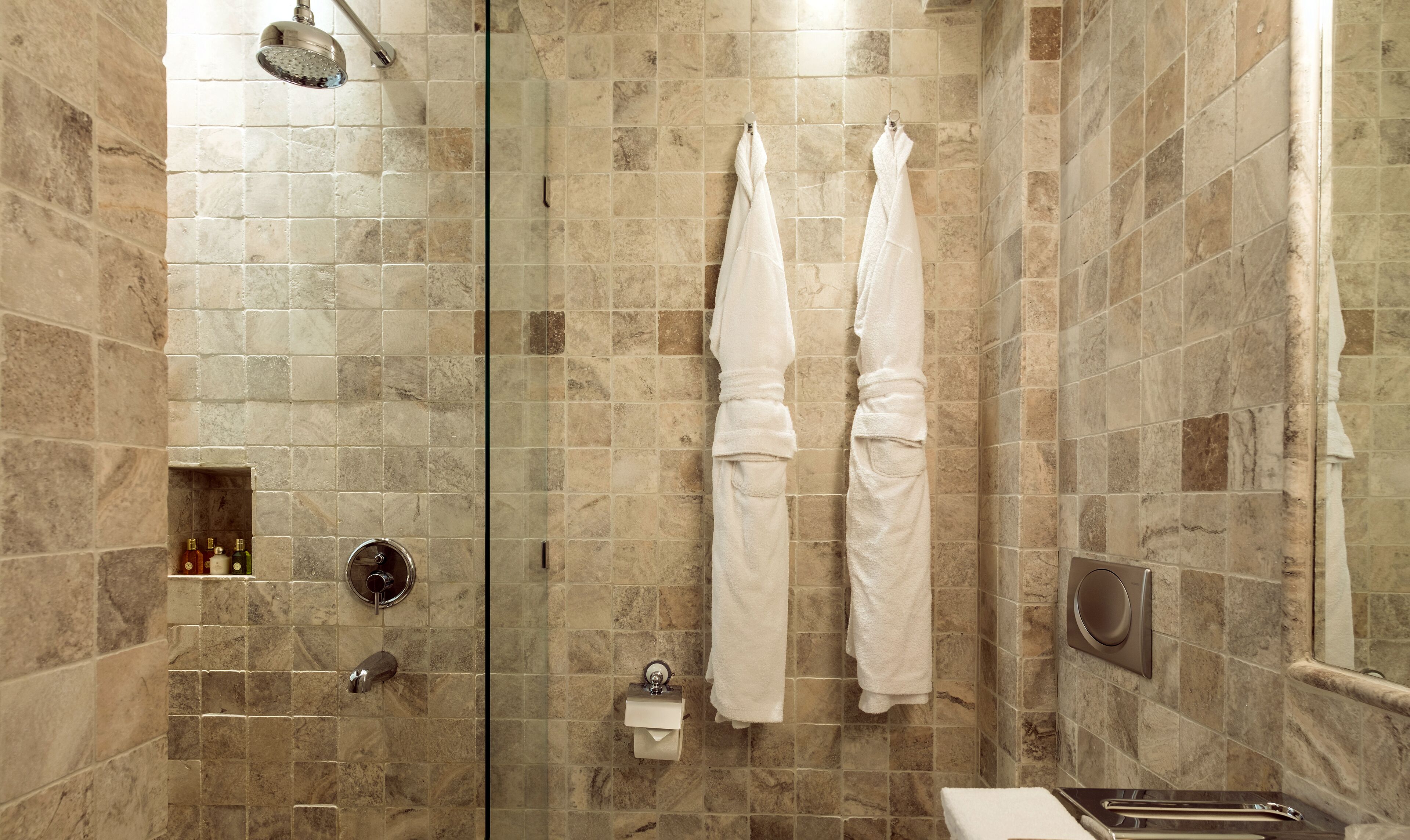 A bathroom with beige tiled walls, a glass shower enclosure, two white towels hanging on hooks, a toilet paper dispenser, and a hand dryer.