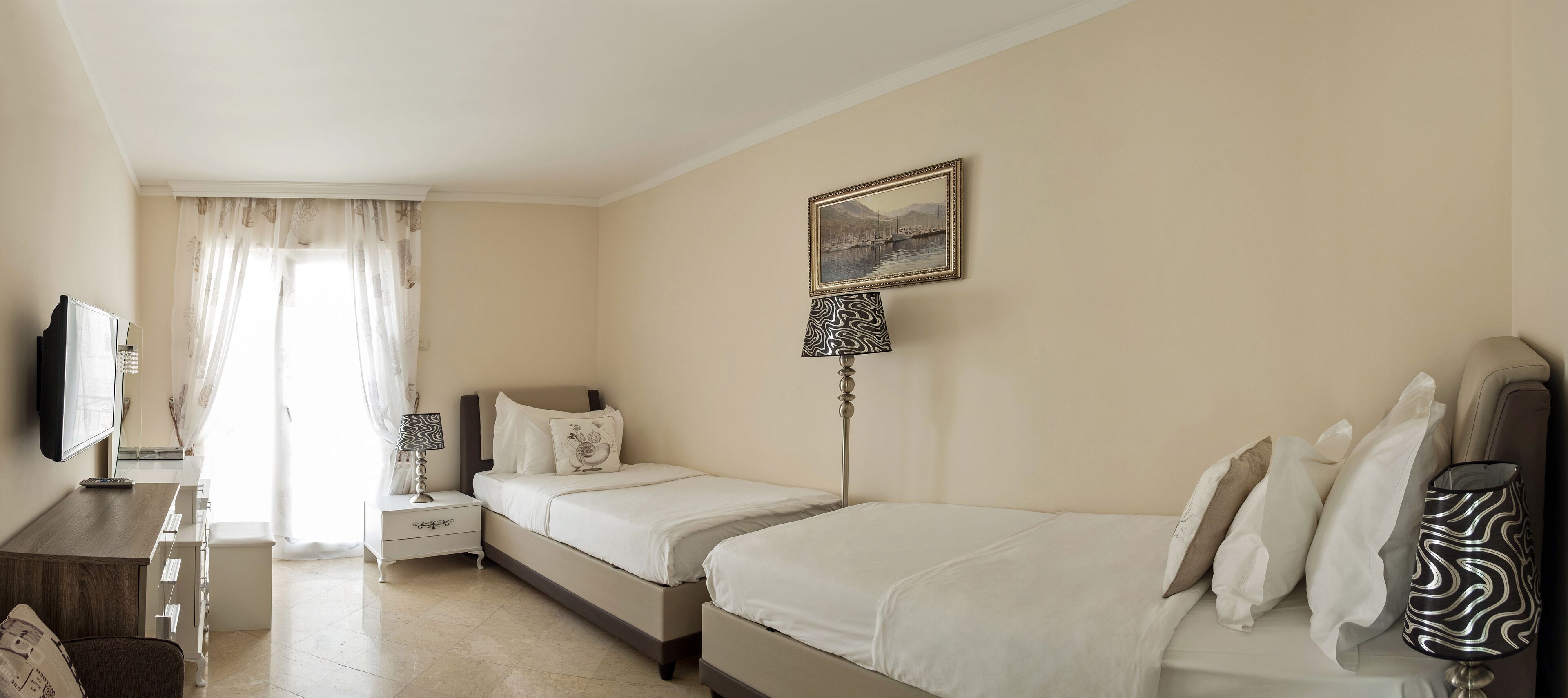 A hotel room with two beds, a desk with a flat-screen TV, a bedside table with a lamp, and a window with sheer curtains.