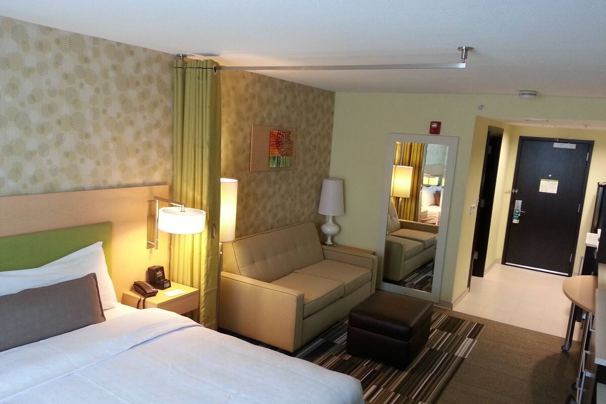 Quarto de hotel com uma cama, um sofá, uma escrivaninha, um espelho e uma porta.
