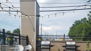 An outdoor patio with a stone fire pit, grilling stations, and string lights. - Home2 Suites by Hilton Rahway, NJ (Rahway)