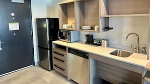 A modern kitchen with a black refrigerator, stainless steel dishwasher, and a microwave. There are wooden shelves above the sink and a coffee maker on the counter. - Home2 Suites by Hilton Rahway, NJ (Rahway)