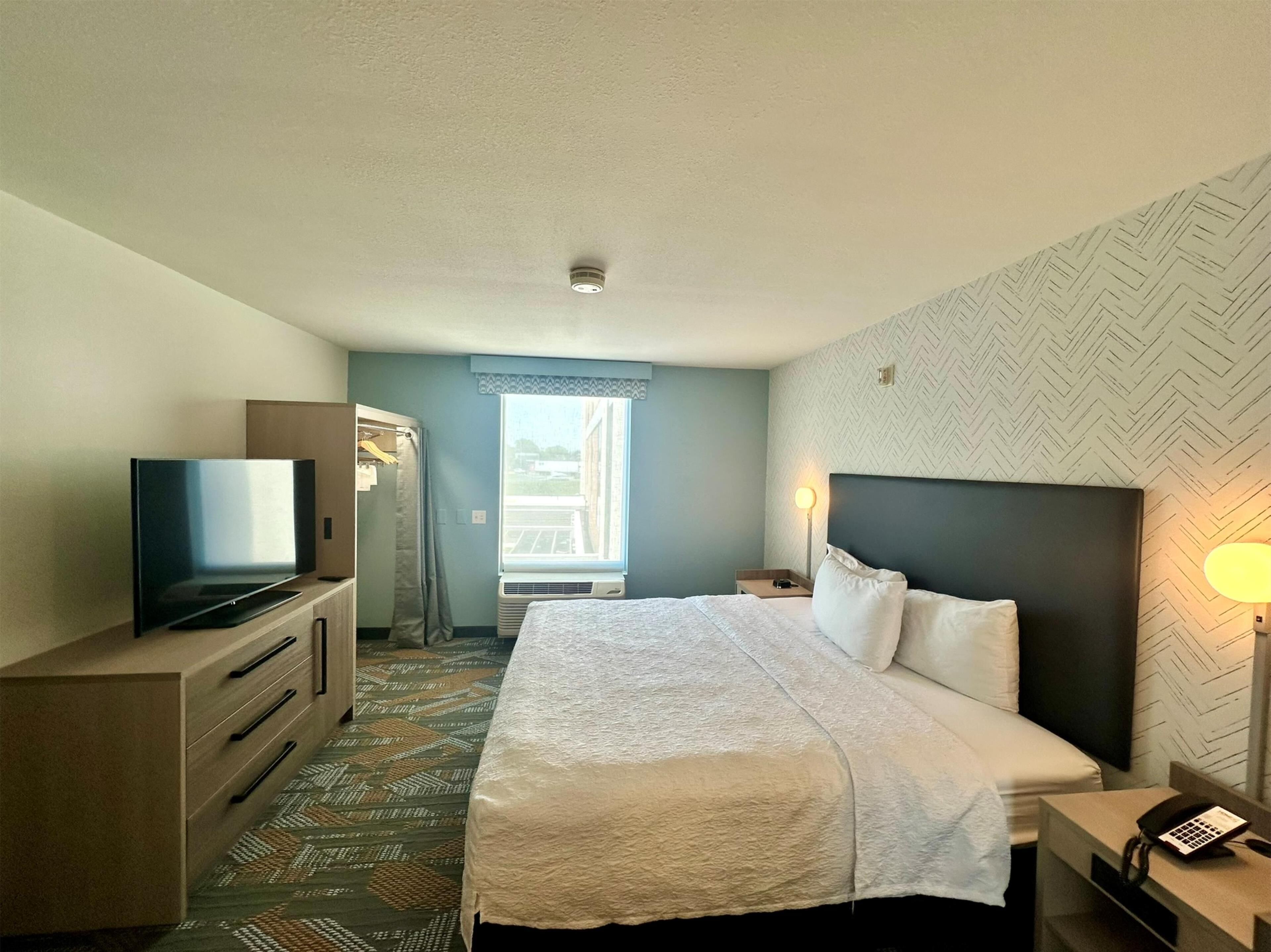 A hotel room with a bed, a TV, a wardrobe, a desk with a lamp, and a window with a view.