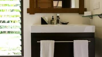 Un baño con un lavamanos blanco, dos toallas colgadas en un perchero, un espejo sobre el lavamanos y una ventana con persianas.