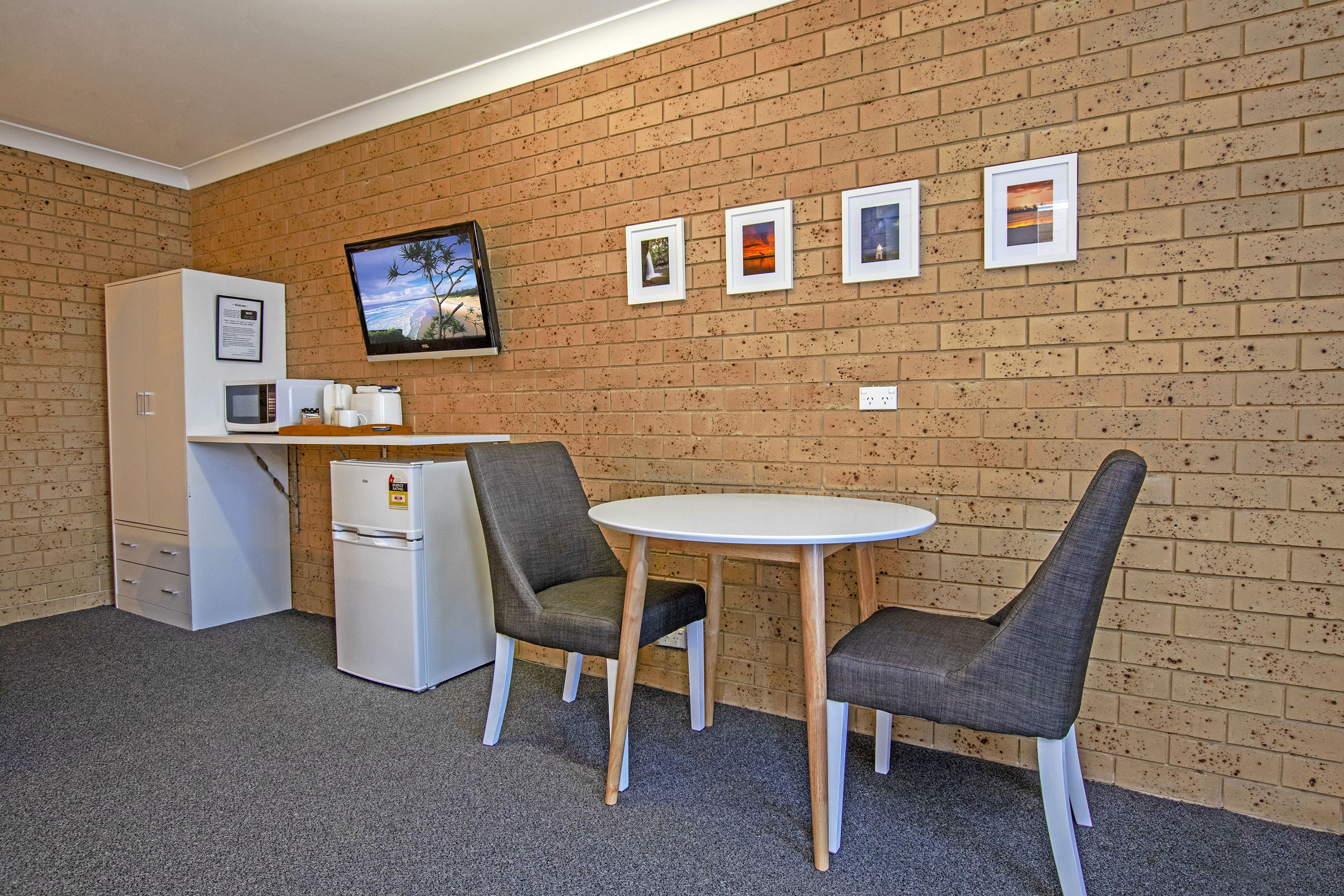 A small kitchenette with a microwave, coffee maker, and refrigerator. There are two chairs and a table in front of a brick wall with four framed pictures.