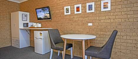 A small kitchenette with a microwave, coffee maker, and refrigerator. There are two chairs and a table in front of a brick wall with four framed pictures.