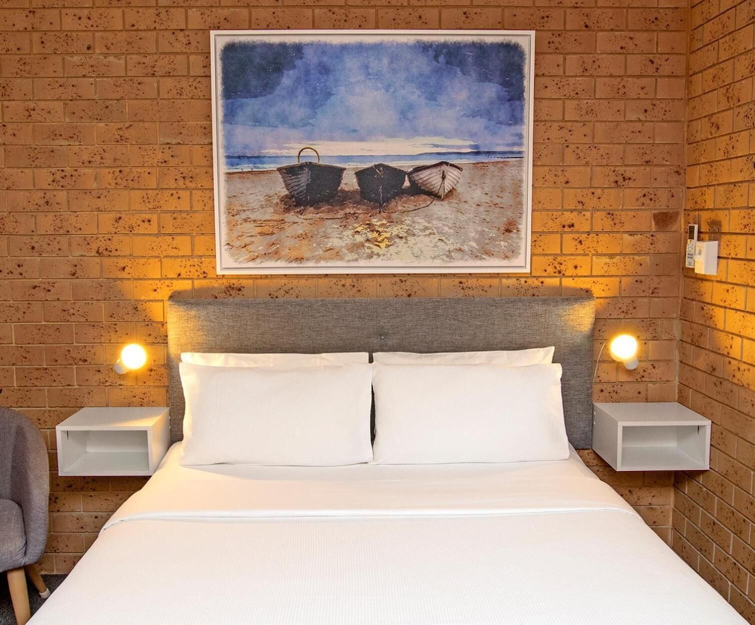 A bedroom with a bed, two bedside tables, a chair, and a painting of boats on a beach.