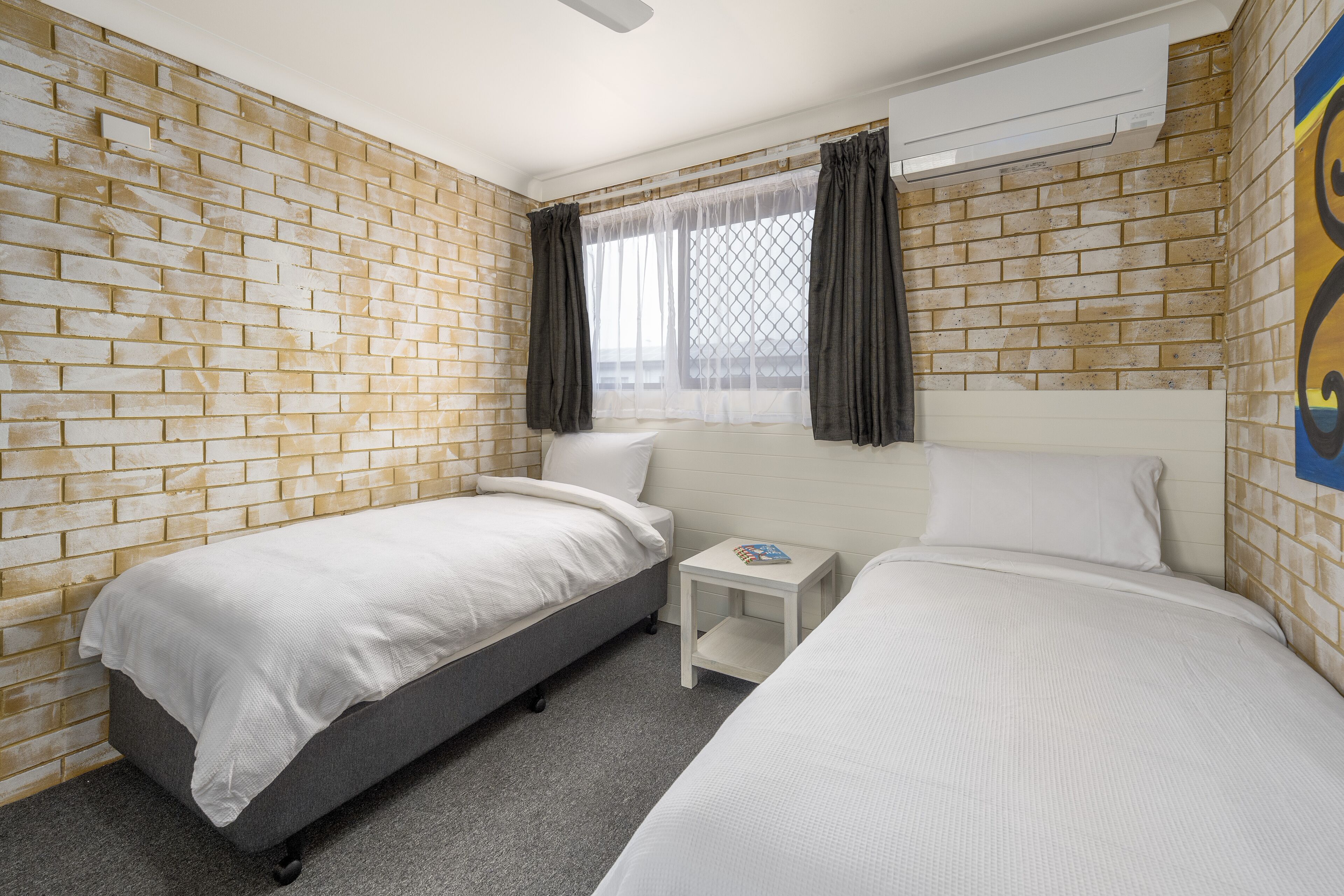 Een hotelkamer met twee bedden, een klein nachtkastje, een raam met gordijnen en een muur met baksteenmotief.