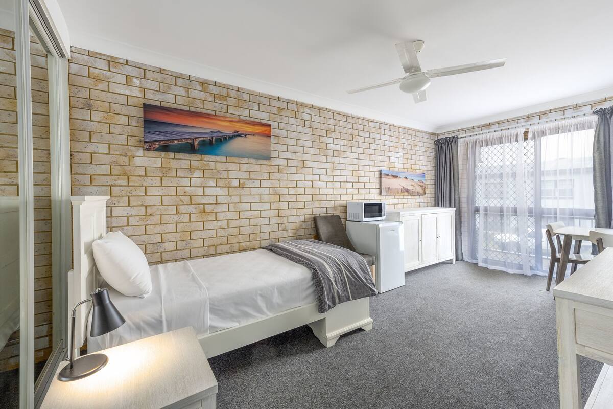 A bedroom with a brick wall, a bed, a desk, a chair, a ceiling fan, and a window with curtains.