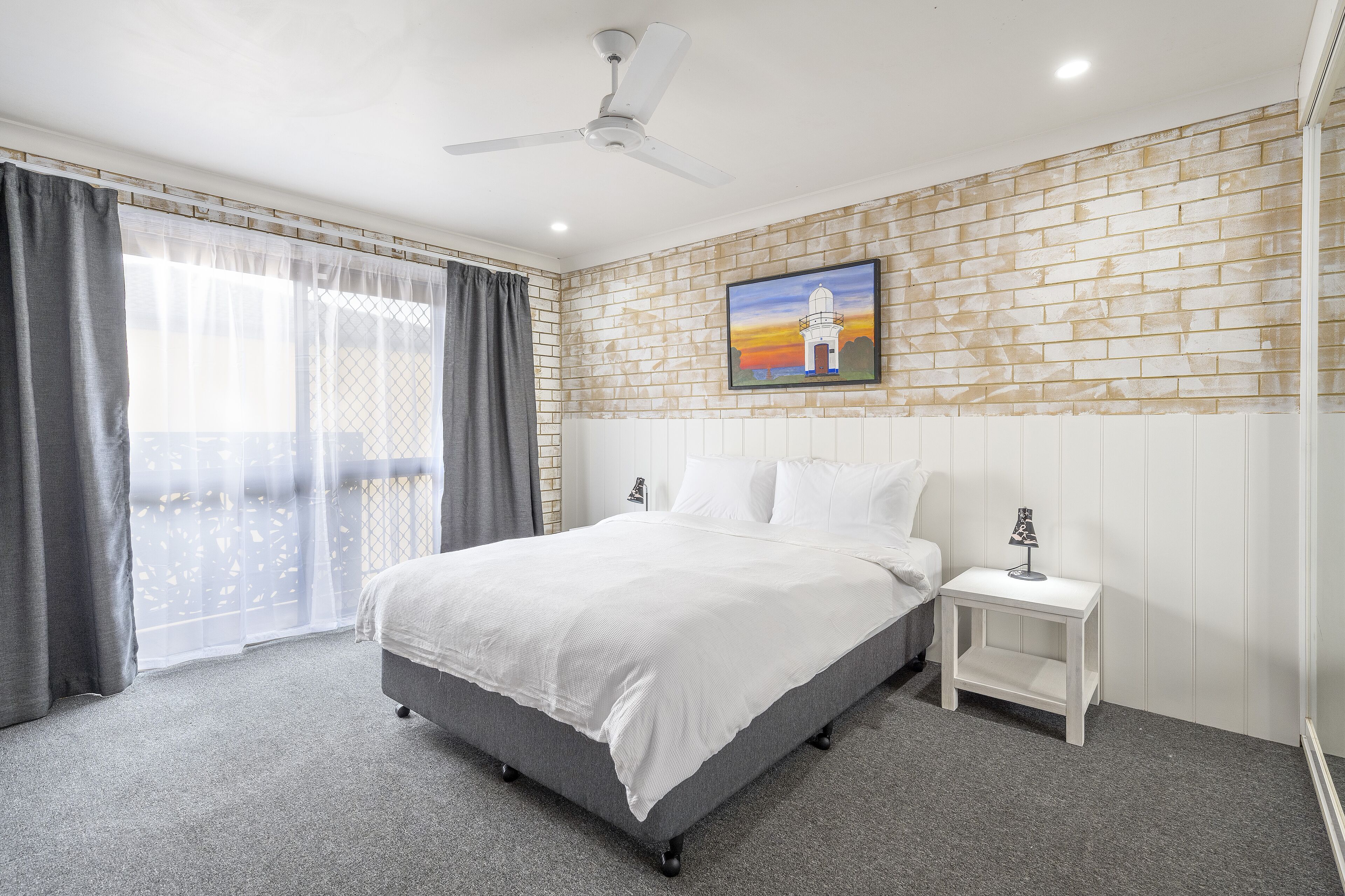 A bedroom with a bed, a nightstand, a painting, and a ceiling fan.