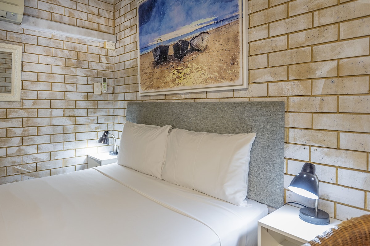 A hotel room with a brick wall, a painting of a beach scene, a bed with white linens, and a nightstand with a lamp.