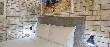 Habitación de hotel con pared de ladrillo, un cuadro de una escena de playa, una cama con sábanas blancas y una mesita de noche con lámpara.
