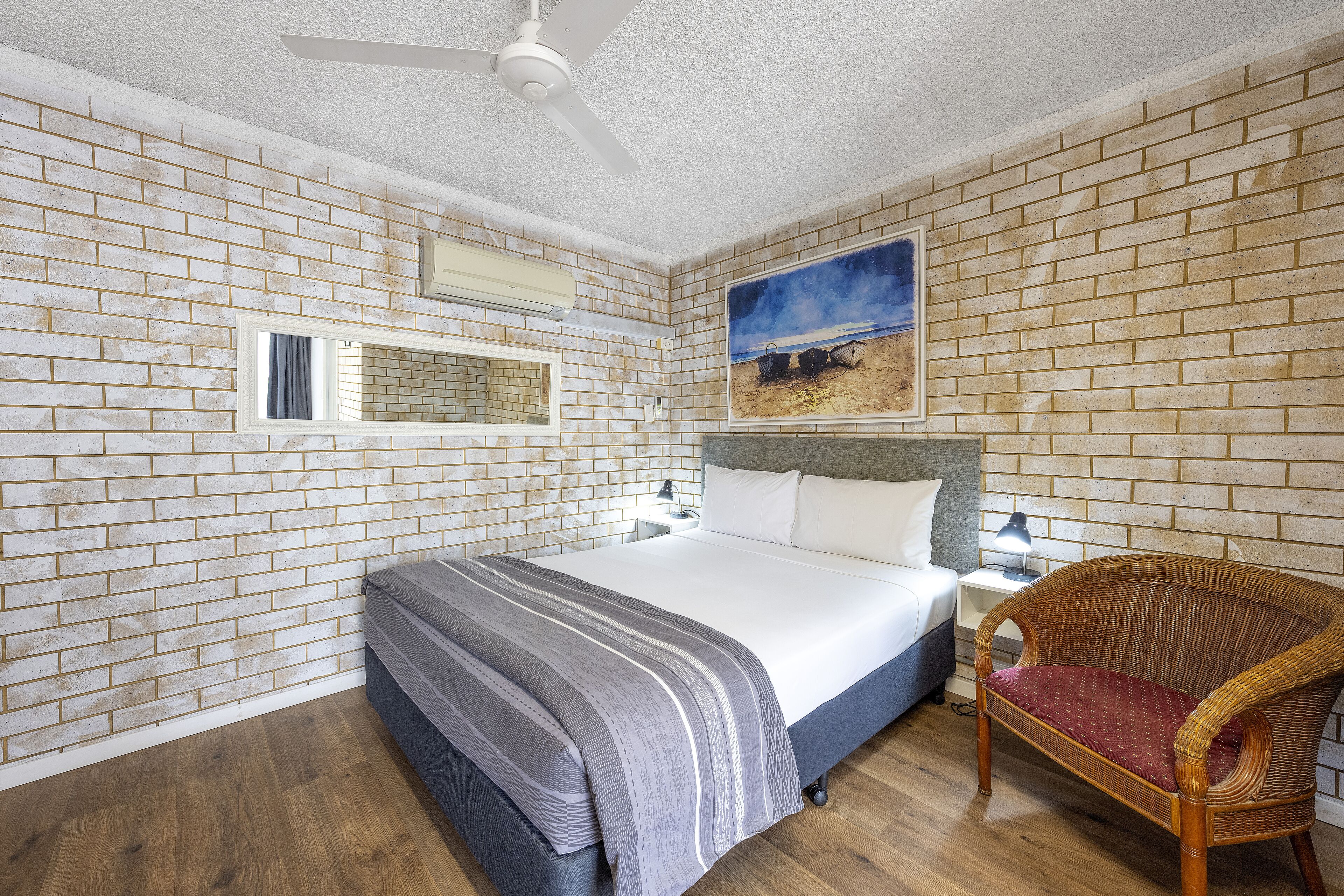 A bedroom with a brick-patterned wall, a bed with a grey and white striped blanket, a wicker chair with a red cushion, and a painting of a beach scene.