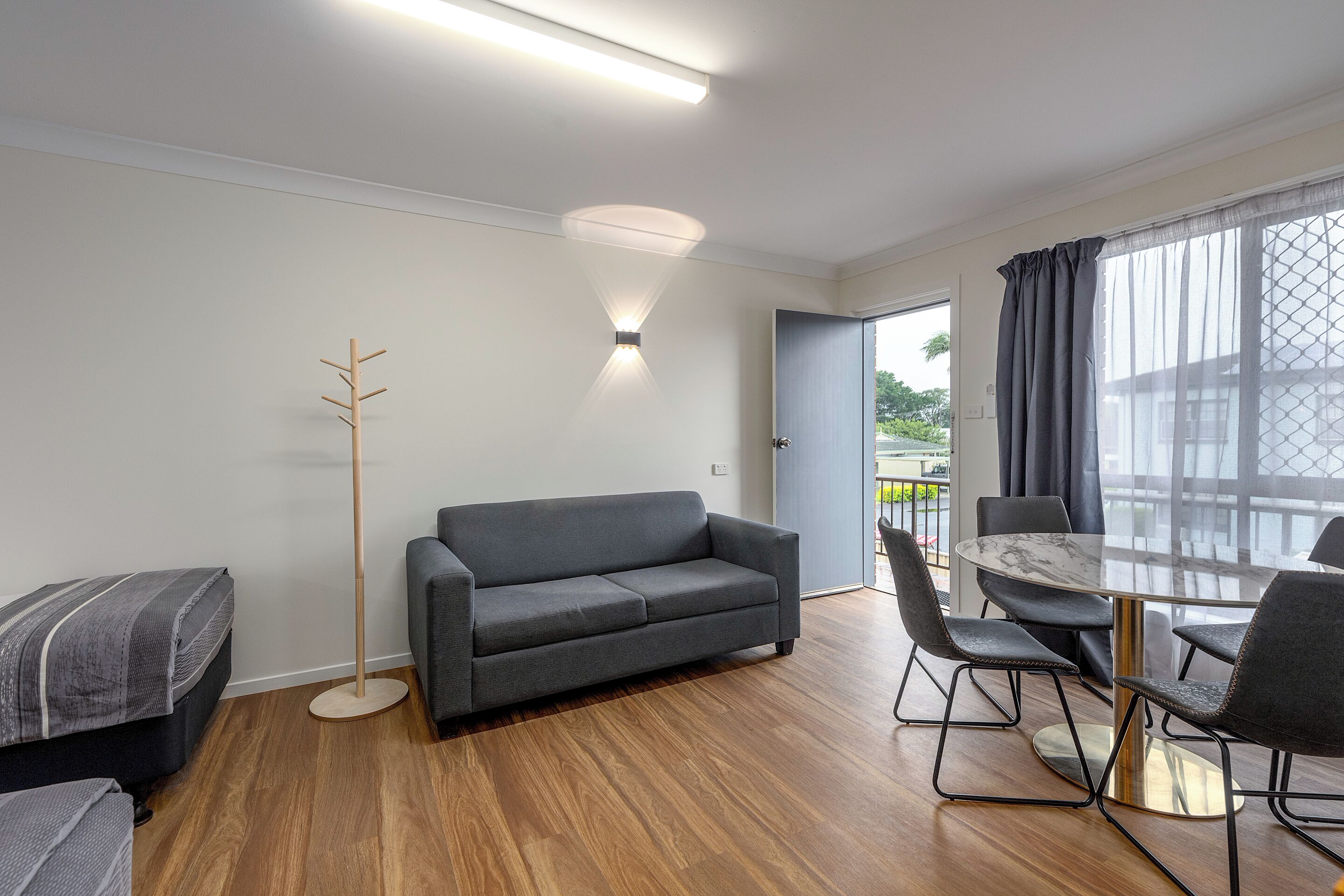 Una habitación moderna con un sofá gris, una lámpara de pie, una mesa de comedor con sillas y vistas a través de un ventanal grande.