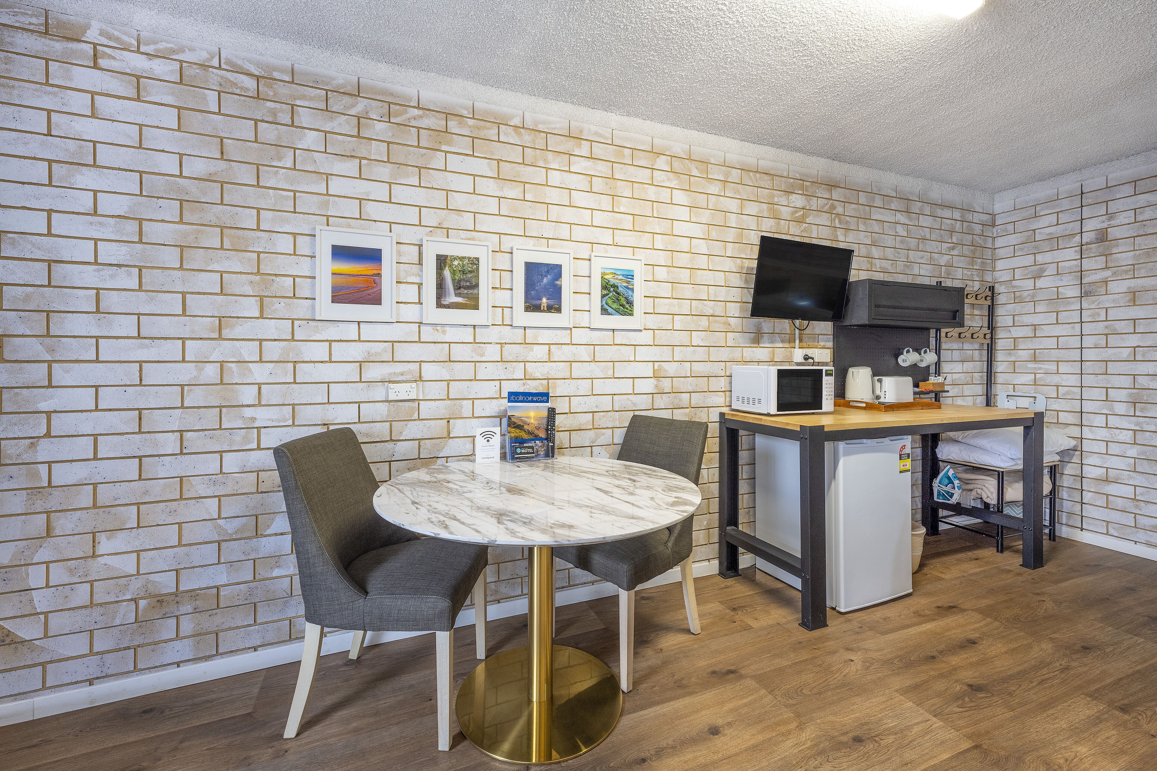 A compact kitchen with a brick wall, a round table with chairs, a microwave, a small fridge, and a TV mounted on the wall.