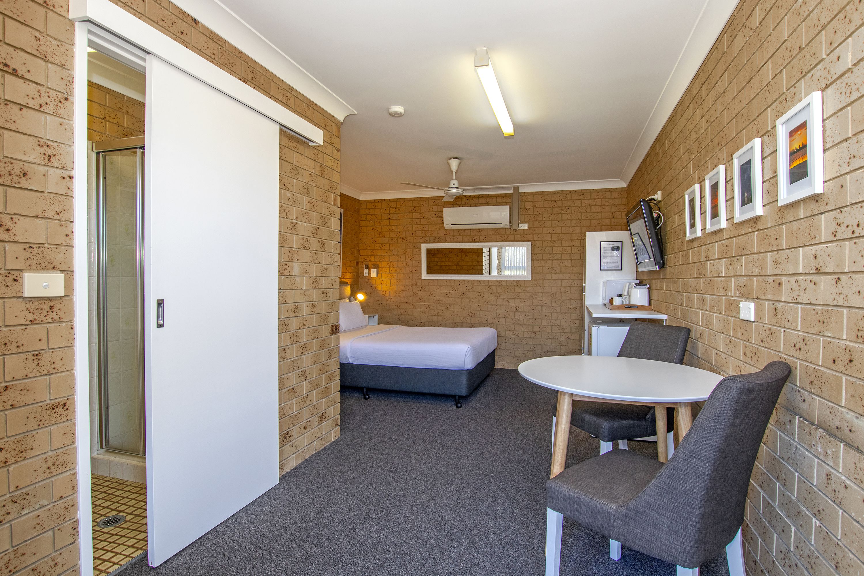 A compact hotel room with a bed, a small desk, a chair, and a shower area.
