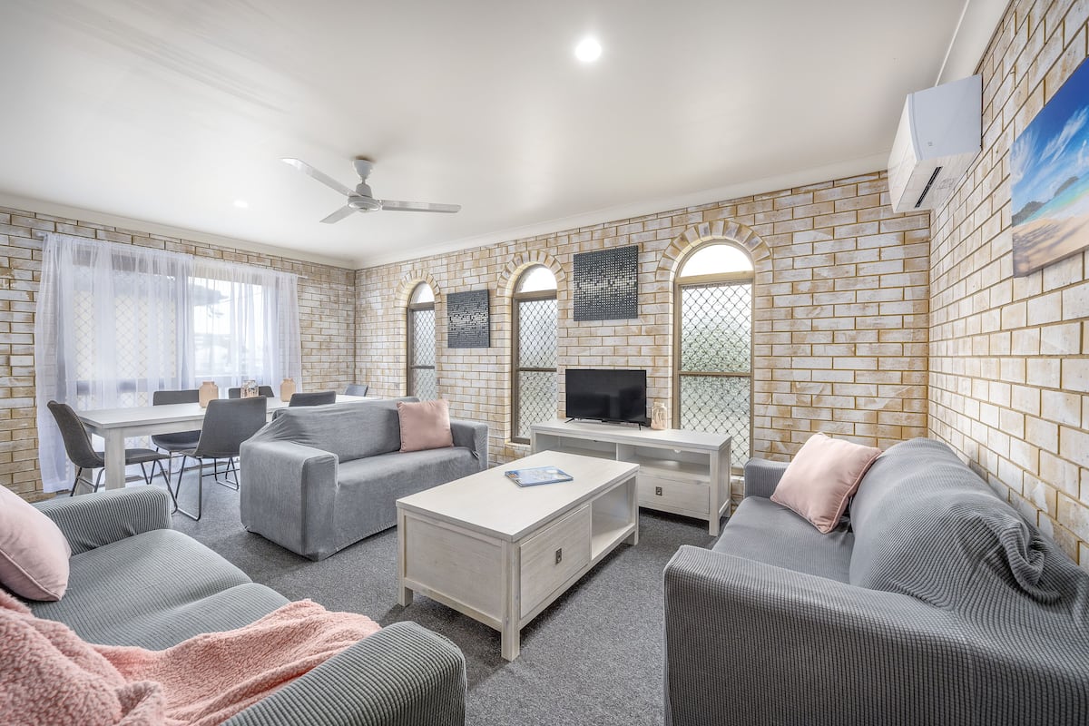 A living room with a sofa, a coffee table, and a TV. There is a dining table with chairs and a window with curtains.