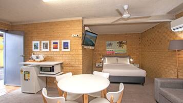 A hotel room with a bed, a desk with a computer, a microwave, a refrigerator, a round table with chairs, and a television mounted on the wall.
