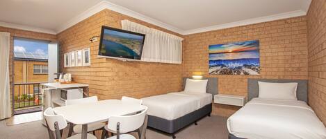 A hotel room with a brick wall, a TV, a desk with chairs, a bed, and a painting of a sunset.
