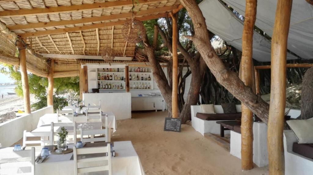 Un área de comedor al aire libre con mesas y sillas de madera, una barra con estantes llenos de botellas y una estructura de techo de paja.