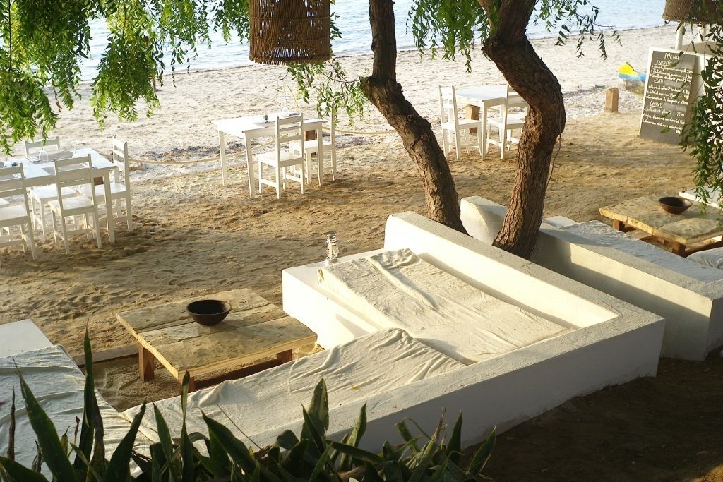 A beachside seating area with white cushioned benches and tables, surrounded by trees and overlooking the beach.