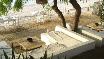 A beachside seating area with white cushioned benches and tables, surrounded by trees and overlooking the beach.