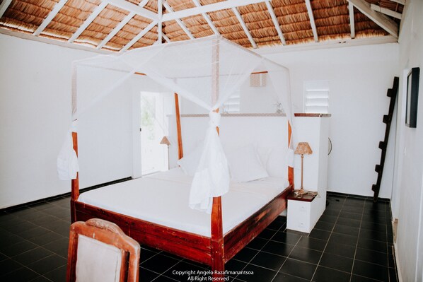 A room with a bed under a mosquito net, a small bedside table with a lamp, and a door with a window.