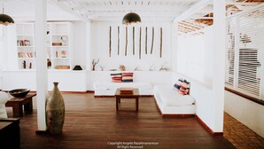 A white interior with a wooden floor, white walls, a white sofa with colorful pillows, a wooden table, and a hanging light fixture. - Princesse du Lagon (Ifaty)