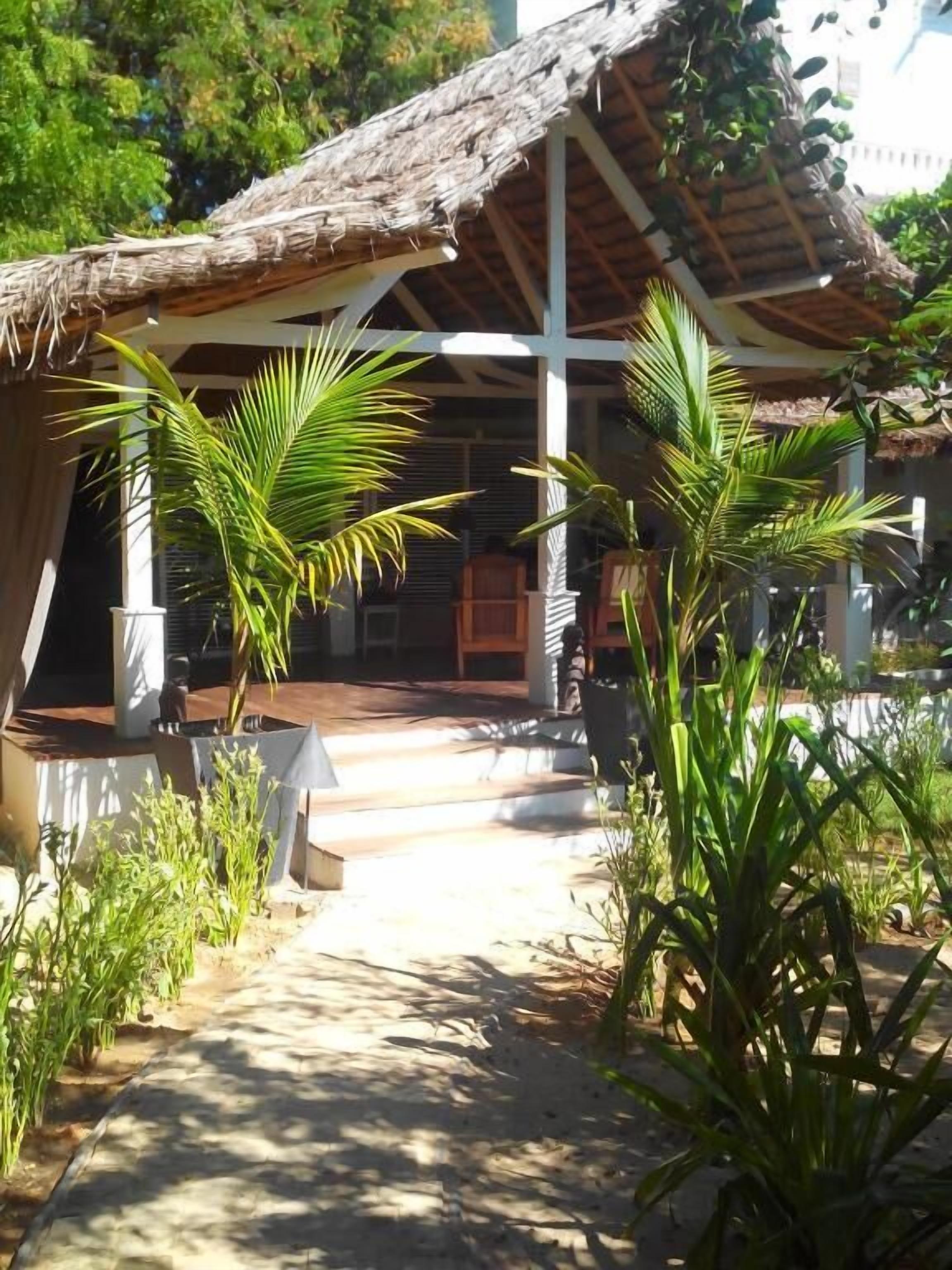 A covered outdoor area with a thatched roof, surrounded by greenery and a pathway leading to a building.