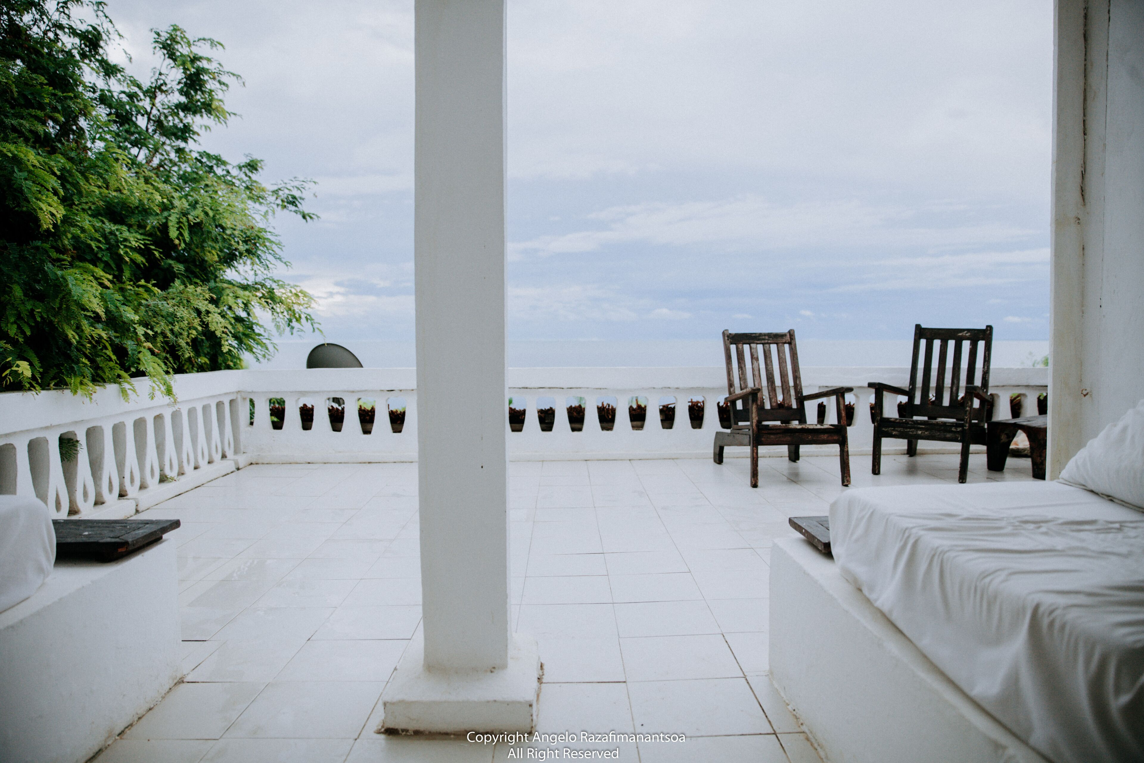 A terrace with white tiled flooring, two wooden chairs, and a bed with white bedding.
