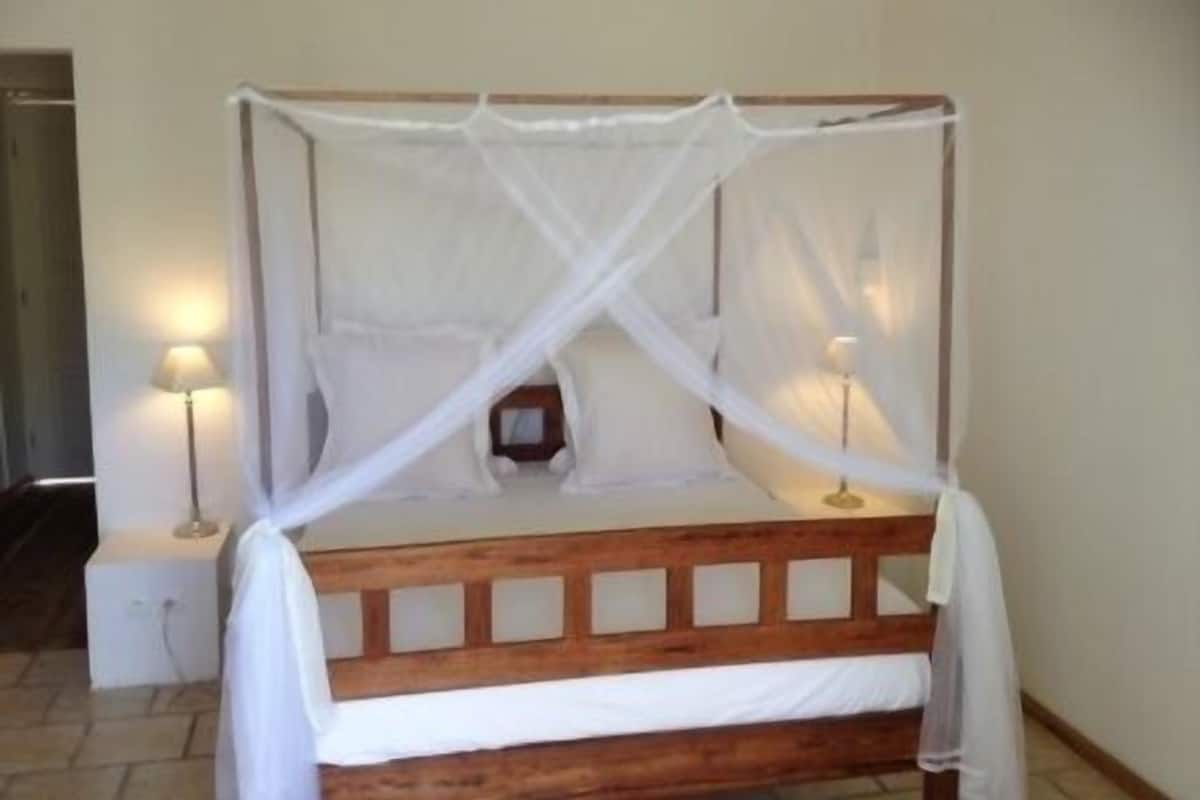 Een bed met houten frame en witte beddengoed, met een hemelbed voorzien van transparante gordijnen.