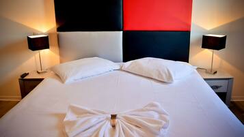 Una cama bien tendida con sábanas blancas, dos almohadas y una toalla dispuestas en el centro. Hay dos mesitas de noche con lámparas a cada lado de la cama.