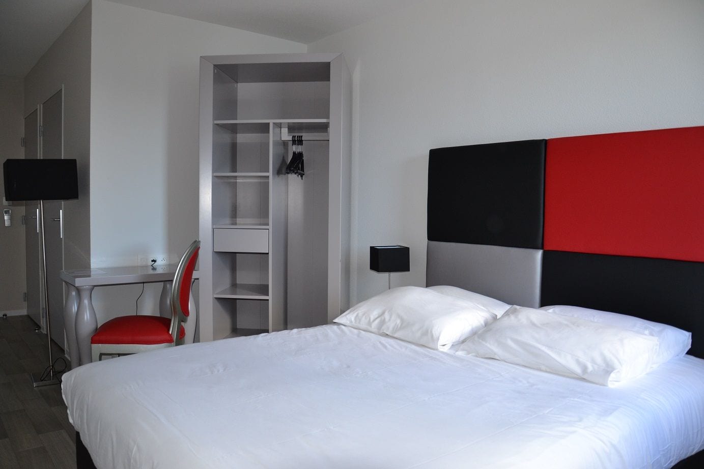 A hotel room with a bed, a desk, a chair, a wardrobe, and a nightstand with a lamp.