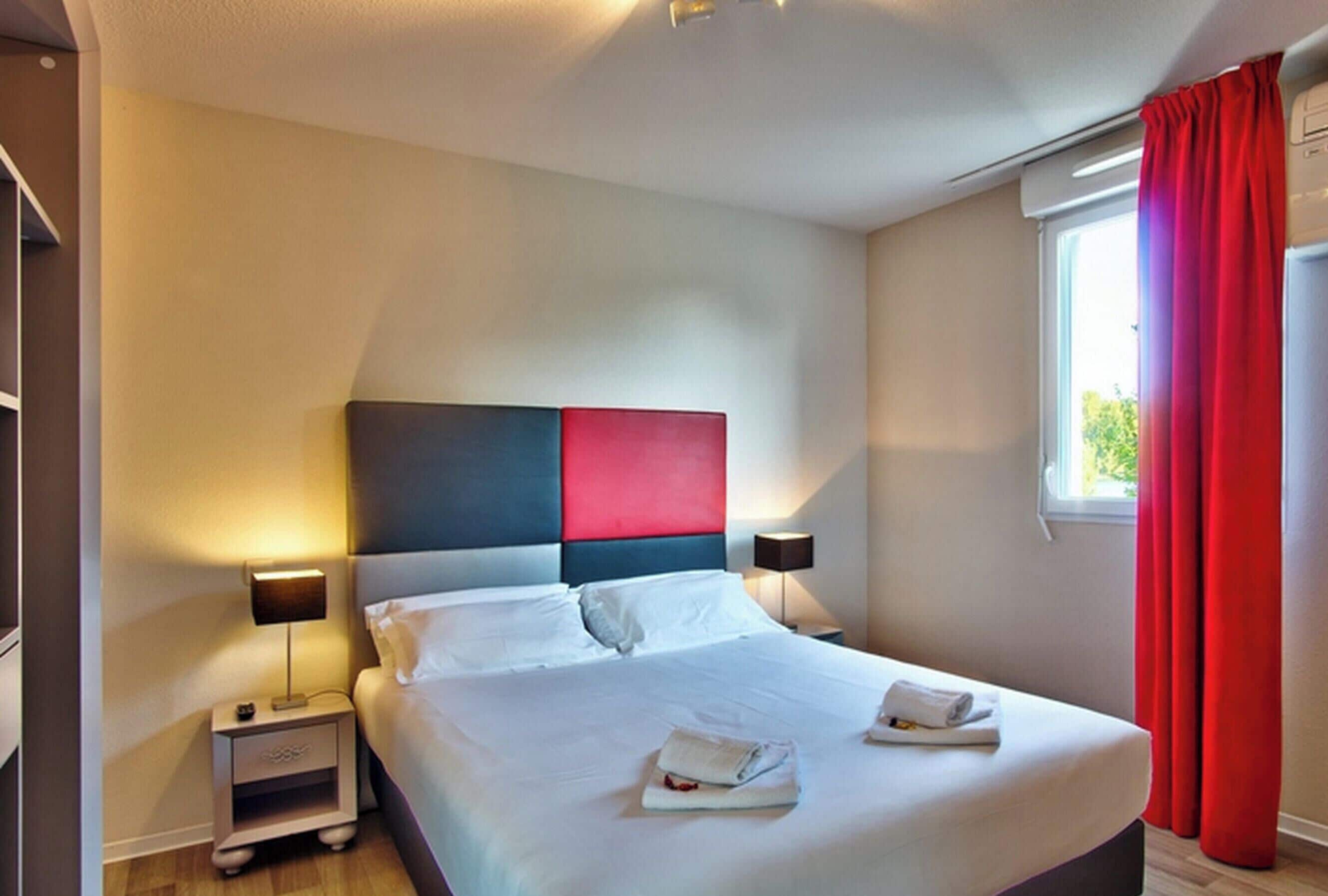 Habitación de hotel con una cama, dos mesitas de noche, un ventanal con cortinas rojas y vistas a zonas verdes.