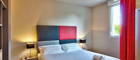 Habitación de hotel con una cama, dos mesitas de noche, un ventanal con cortinas rojas y vistas a zonas verdes.