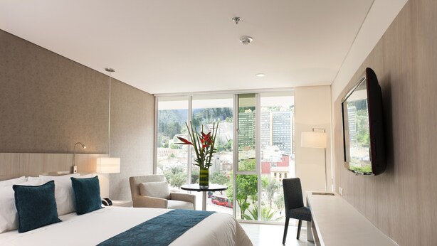 Quarto de hotel moderno com uma cama grande, uma escrivaninha, uma cadeira, uma televisão e vista para a cidade.