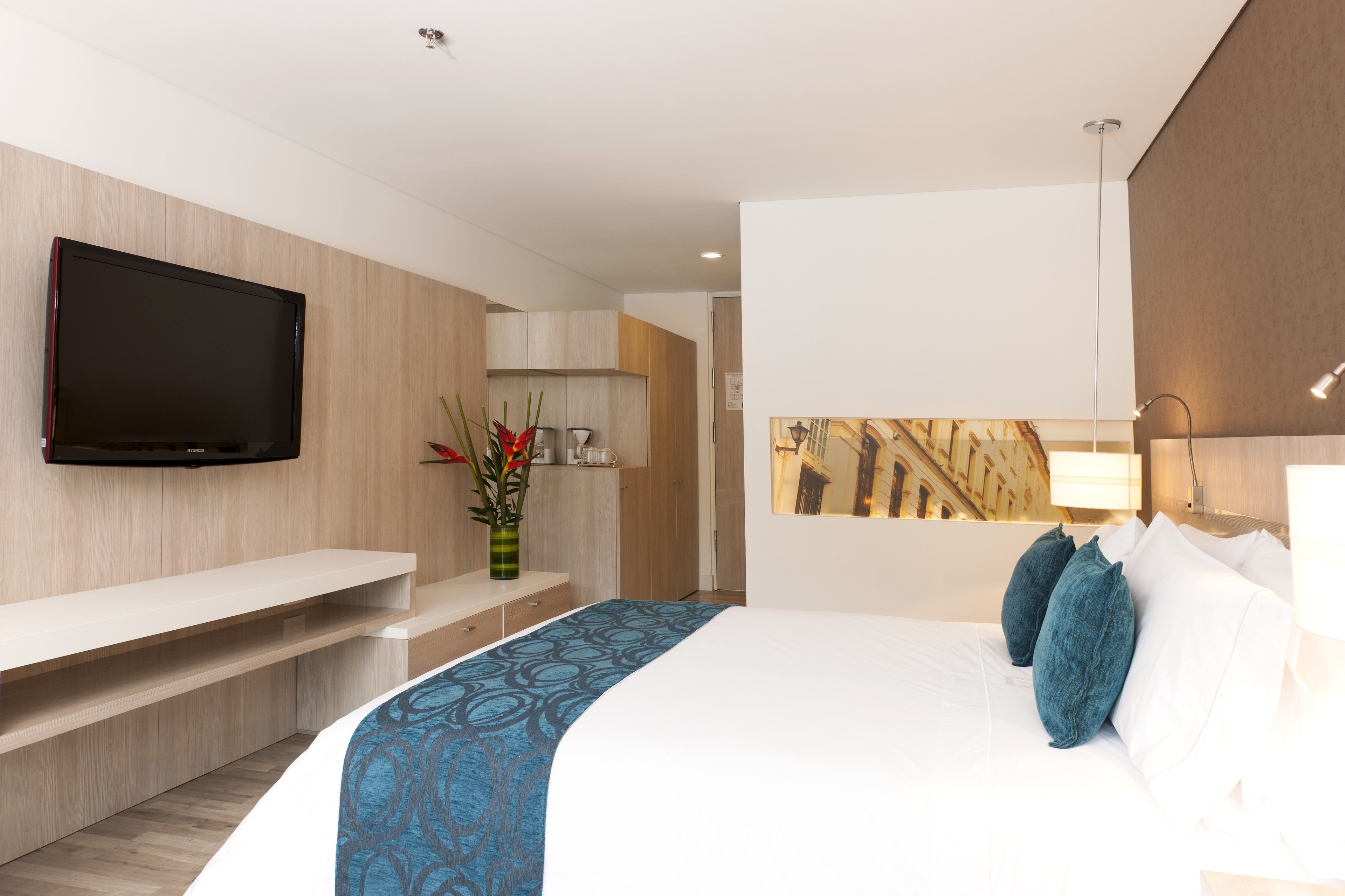 A modern hotel room with a flat-screen TV mounted on the wall, a bed with blue and white bedding, a bedside table, and a small desk area.