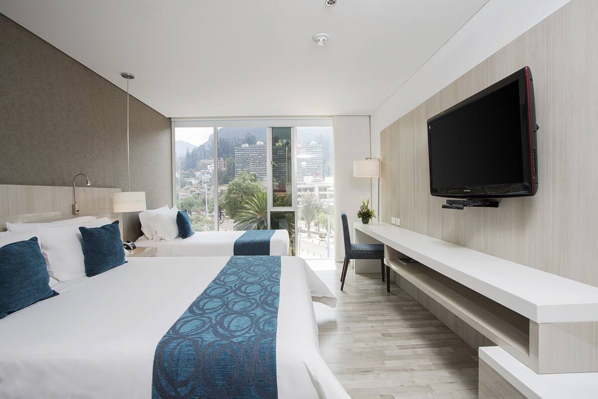 A hotel room with two beds, a TV, a desk, and a large window with a city view.