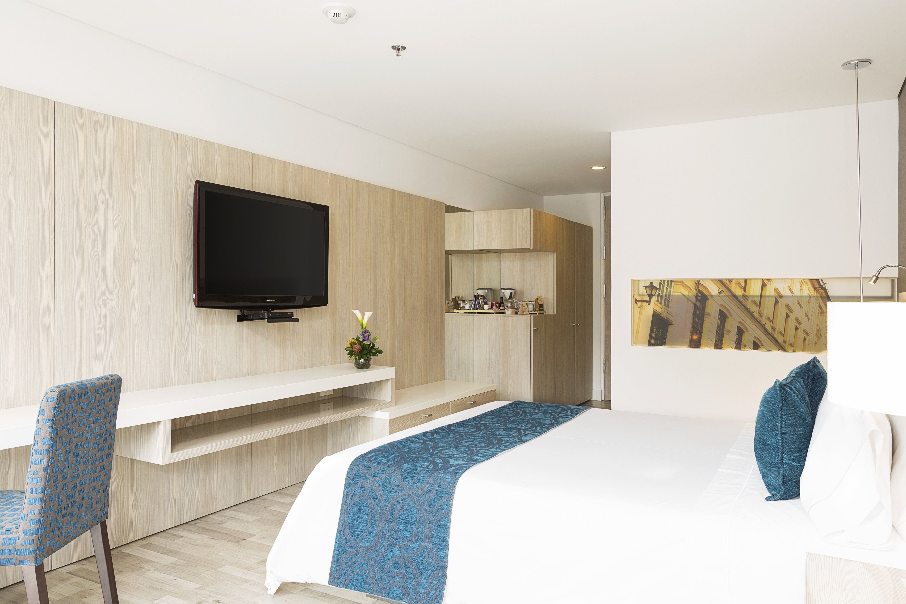 Ein modernes Hotelzimmer mit einem großen Bett, einem an der Wand montierten Fernseher, einer Sitzecke mit einem Sessel und einem kleinen Tisch mit Blumenvase.