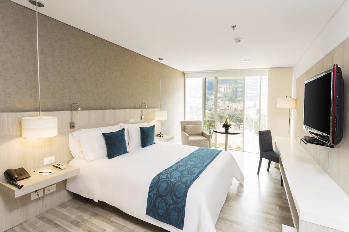 A hotel room with a large bed, bedside tables, a desk with a chair, a TV, and a view of the city through large windows.