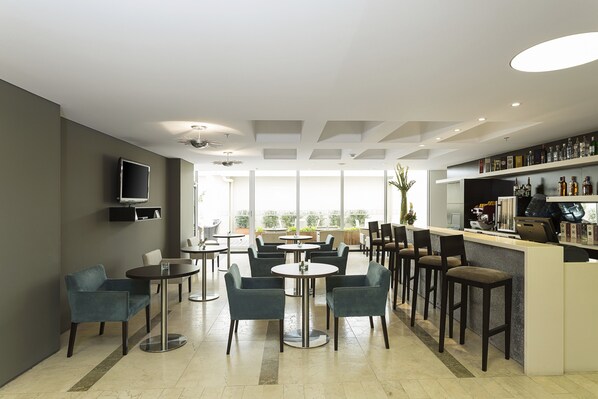 A modern bar area with round tables and chairs, a bar counter with a variety of bottles, and a large window providing a view of the outdoors.