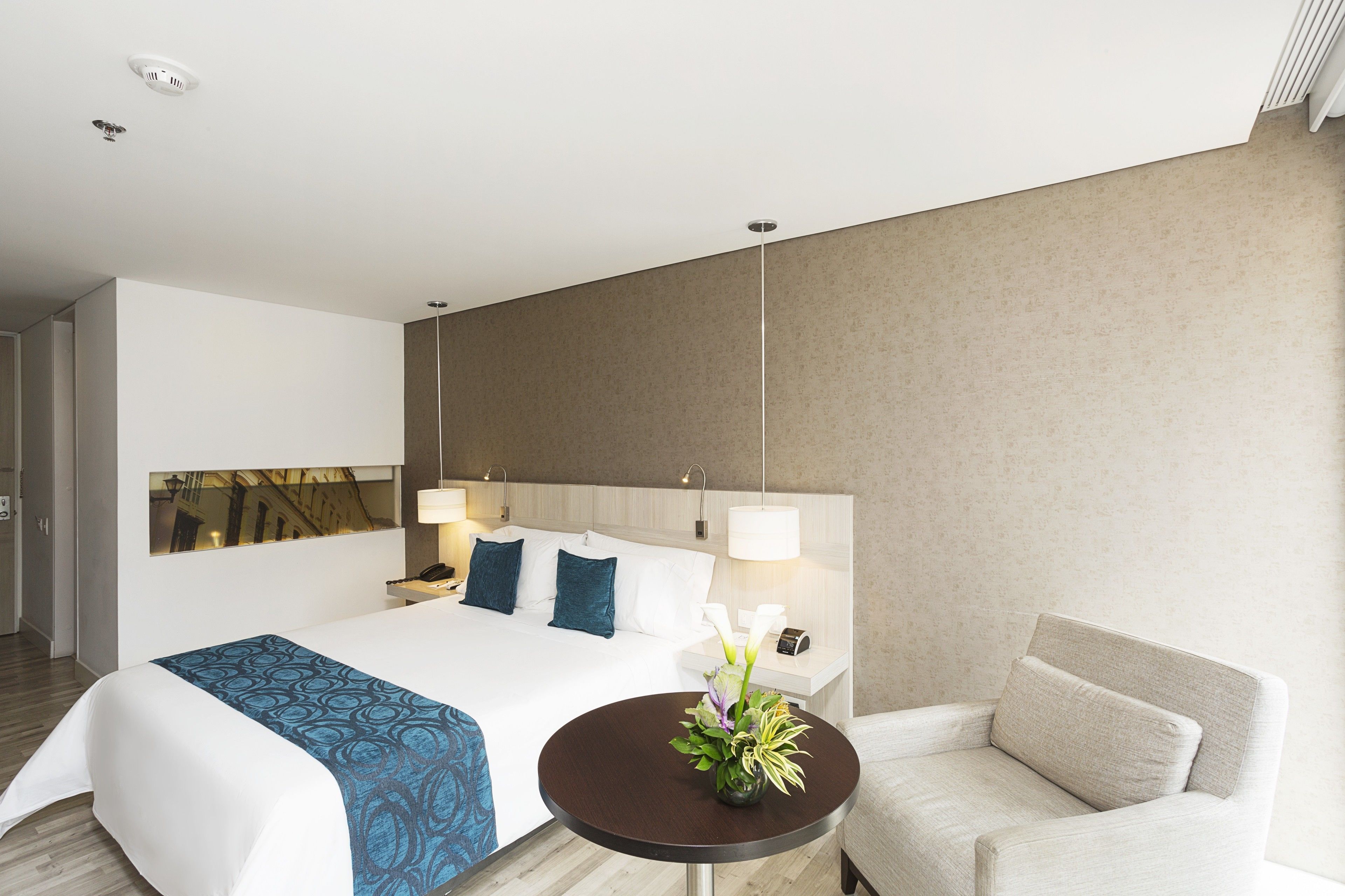 Una habitación de hotel moderna con una cama grande, lámparas de noche, una mesita redonda con un florero y una silla.