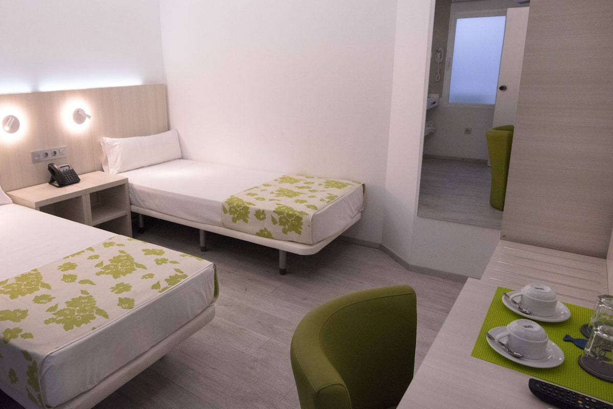 Quarto de hotel com duas camas, uma escrivaninha com telefone, uma cadeira e uma mesa com chávena e pires.