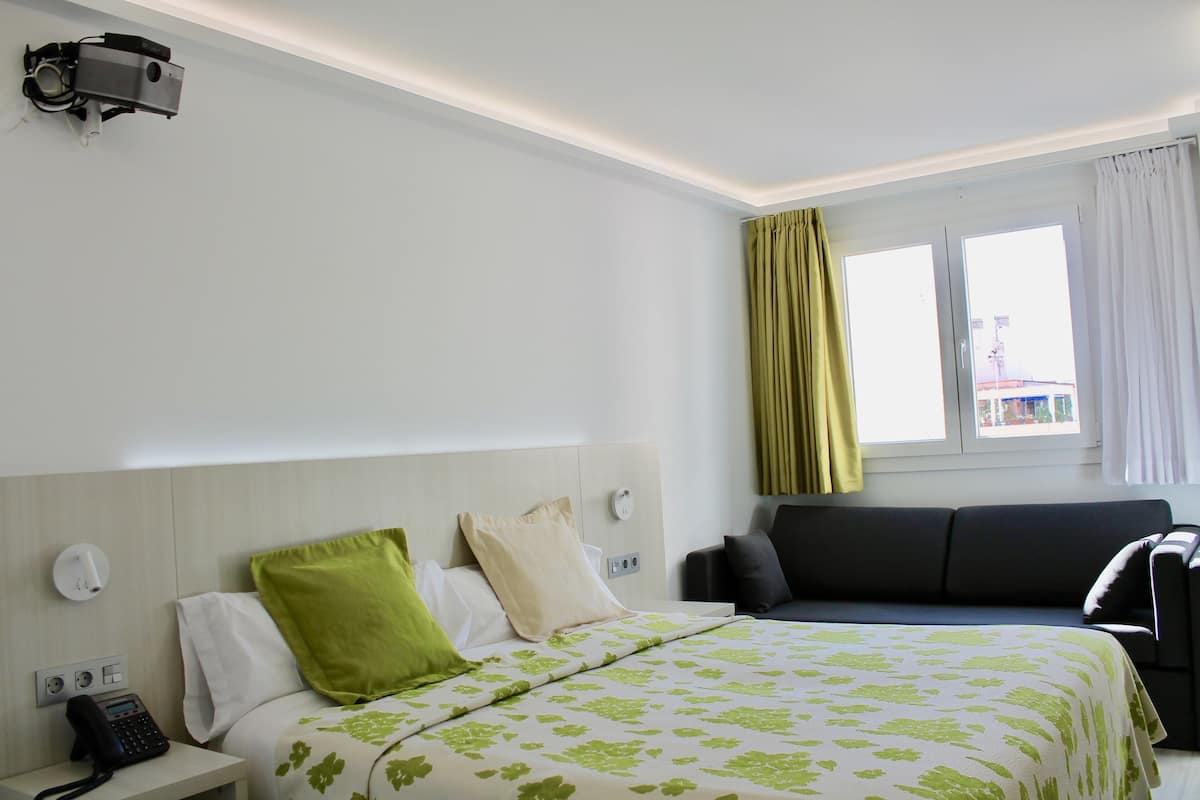 Quarto de hotel com cama, mesa de cabeceira com telefone, projetor fixado na parede, janela com cortinas e um sofá.