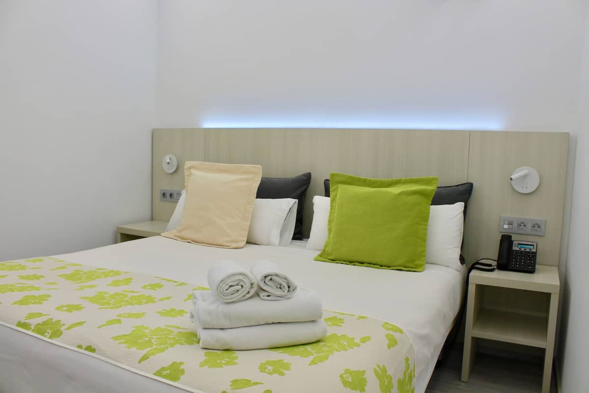 Quarto de hotel com uma cama bem arrumada, mesa de cabeceira com telefone e luminária na parede.