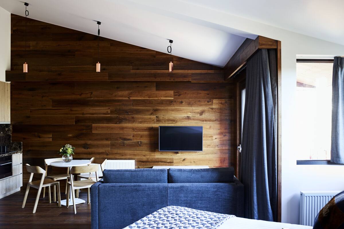 A modern hotel room with a wooden wall, a flat-screen TV, a sofa, a dining area with chairs, and a window with curtains.